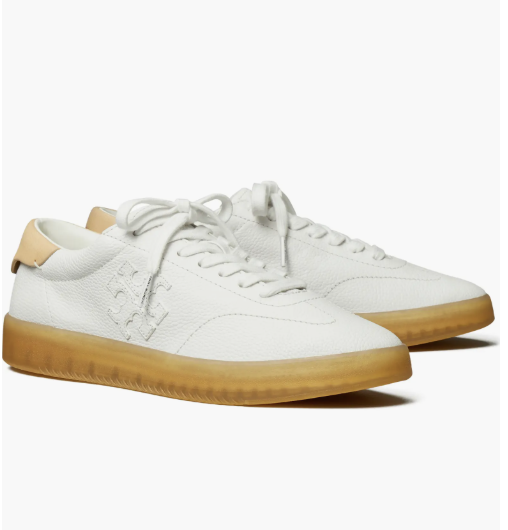 A product shot of the Tory Burch Phoenix Sneaker, a white pebbled leather low-top sneaker featuring an embossed logo, tan heel tab, and a classic gum rubber sole.