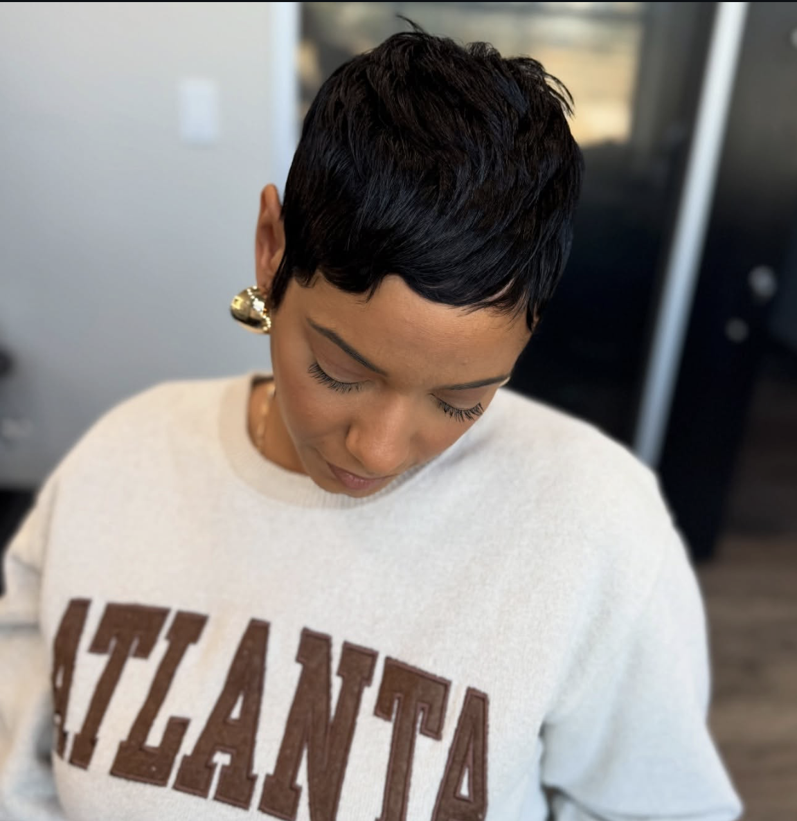 A close-up view of a woman with a sharp, textured black pixie cut wearing a gray Atlanta sweatshirt and gold earrings.