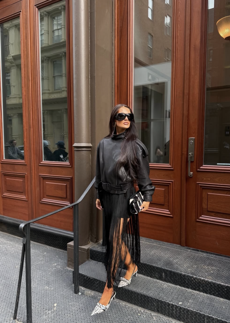 A woman wearing a black leather moto jacket and a black midi skirt with long, dramatic fringe, paired with pointed-toe patterned heels.