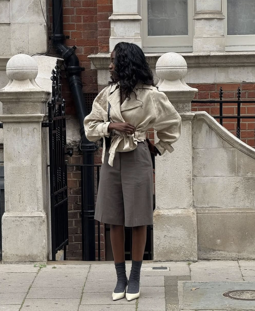 A woman standing outside a brick building wearing a beige belted wrap jacket, dark grey tailored bermuda shorts, and charcoal grey ribbed crew socks.