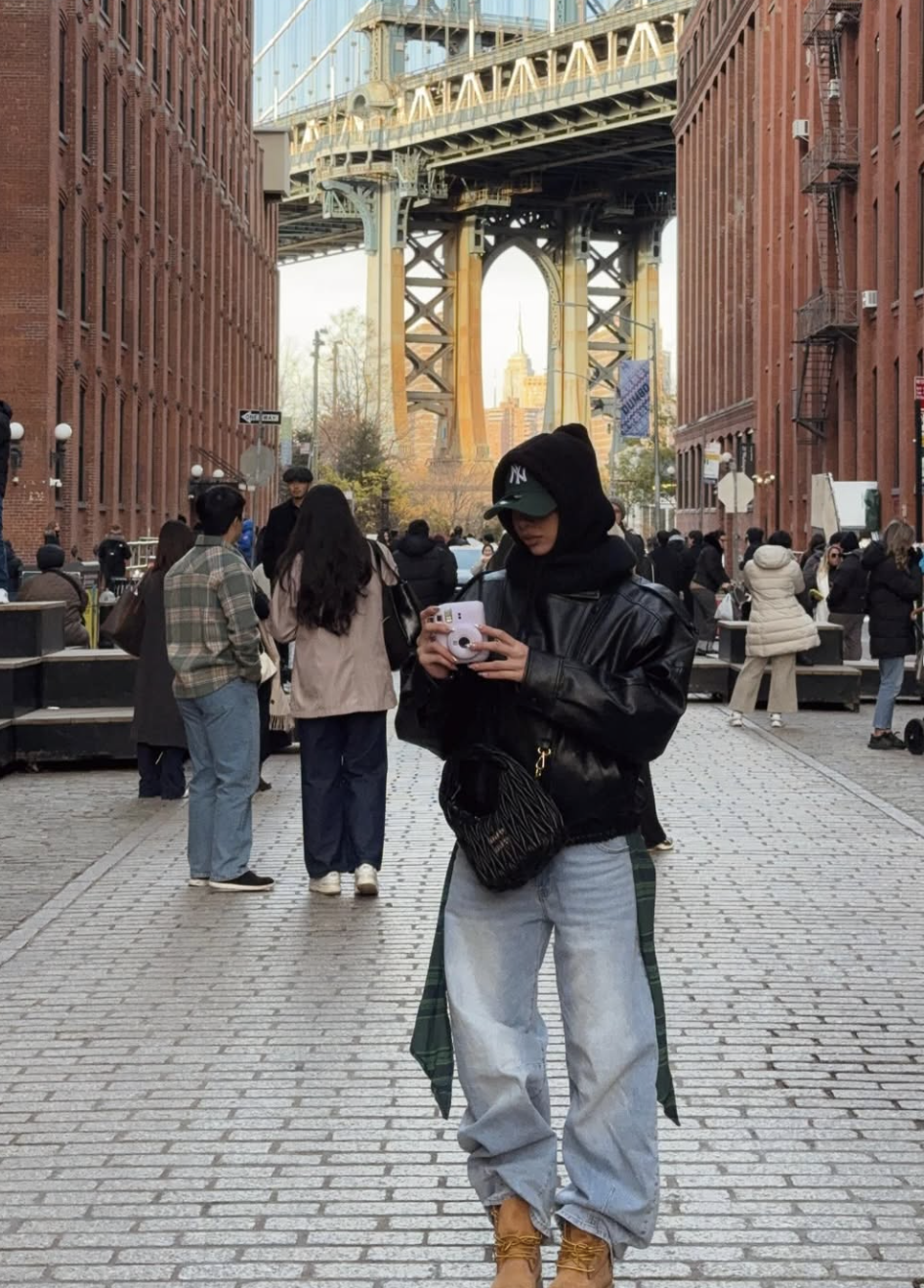 A woman wearing light-wash wide-leg jeans cuffed over tan utility boots, paired with a black leather jacket and knit beanie in an architectural city setting.