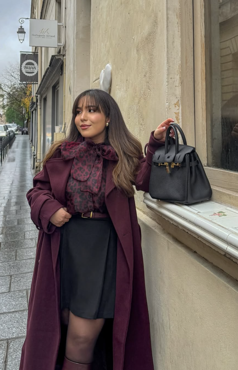 A woman wearing a long burgundy coat over a leopard print pussy-bow blouse and a black mini skirt, holding a black handbag