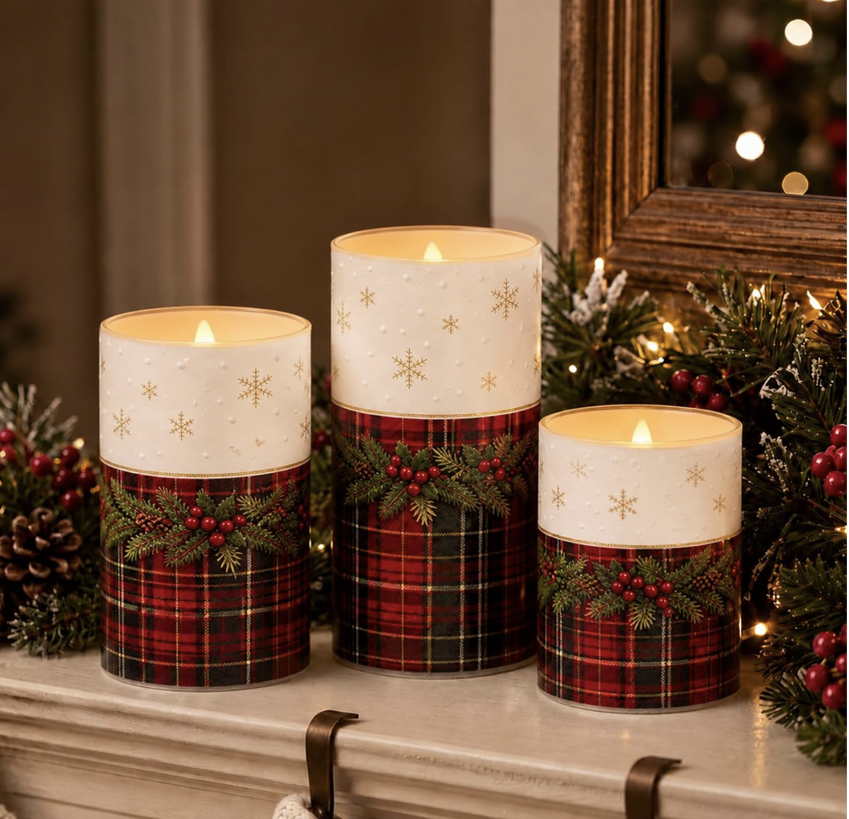 Three red tartan flameless candles with gold snowflakes and holly on a mantel lined with evergreen, pinecones and red berries.