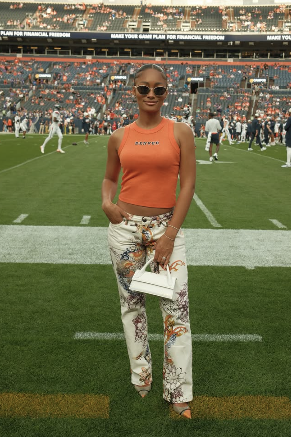 Woman wearing an orange Denver tank with cream printed trousers and a small white top-handle bag at a football game.