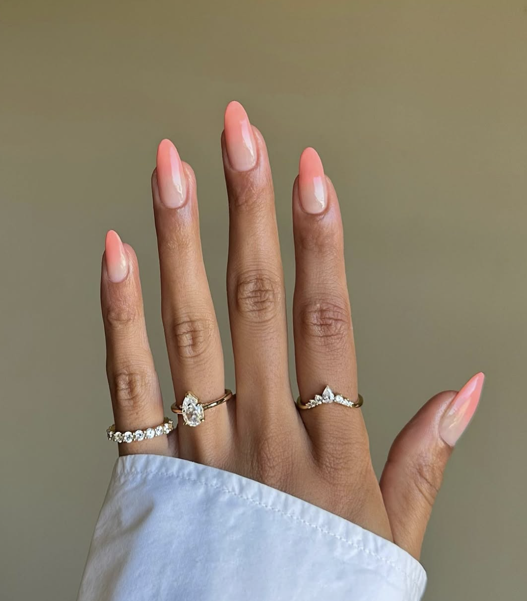 Long almond-shaped gel nails with a soft peach-to-nude ombre gradient and gold ring accessories.