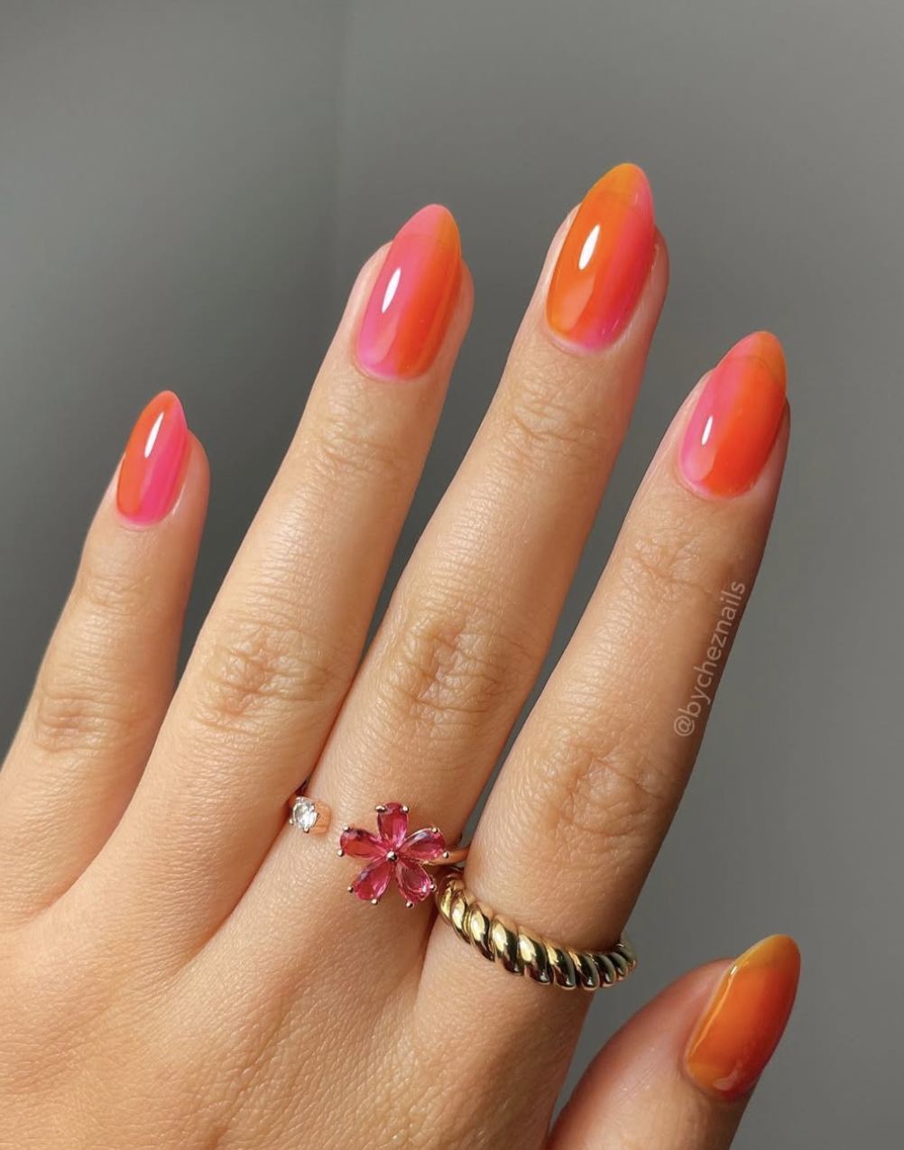 Close-up of a hand with long almond-shaped nails showcasing a vibrant summer gradient manicure that transitions from a bright neon pink at the base to a juicy tangerine orange at the tips, styled with a gold ring featuring a pink flower charm.