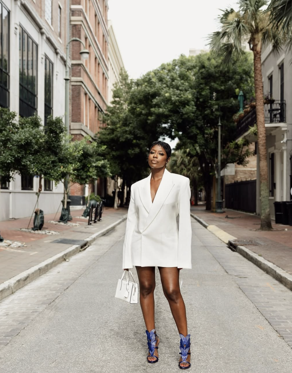 A woman with short hair standing on a city street wearing a white tailored blazer dress paired with vibrant blue strappy heeled sandals.