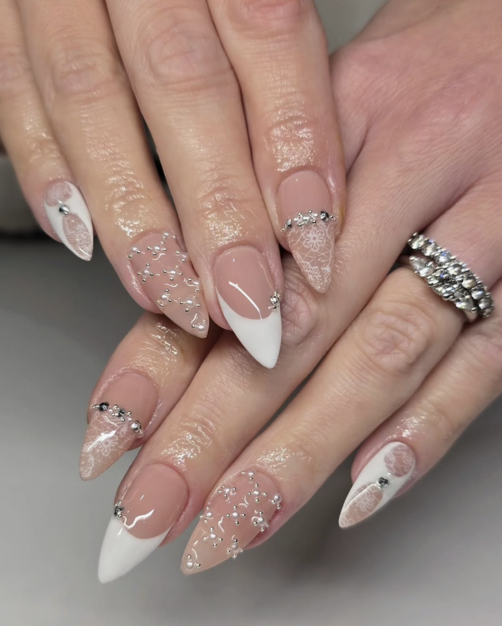 Long almond and stiletto nails with a sheer nude base featuring a mix of solid white French tips, intricate white lace patterns, and 3D silver rhinestone embellishments.