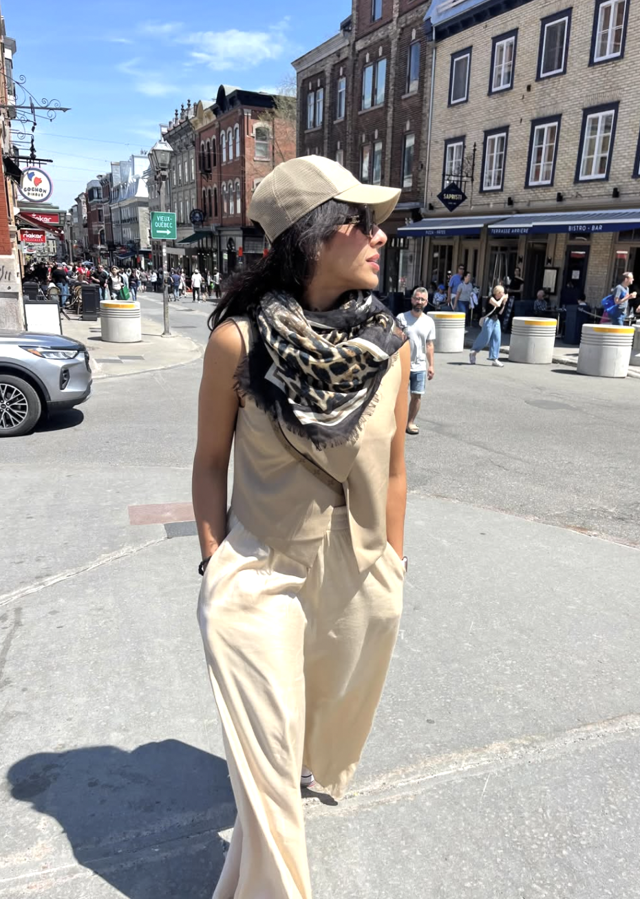 A street style shot of a woman wearing a neutral leopard print scarf styled as a full front drape over a tailored beige vest and matching high-waisted trousers, completed with a tan baseball cap.