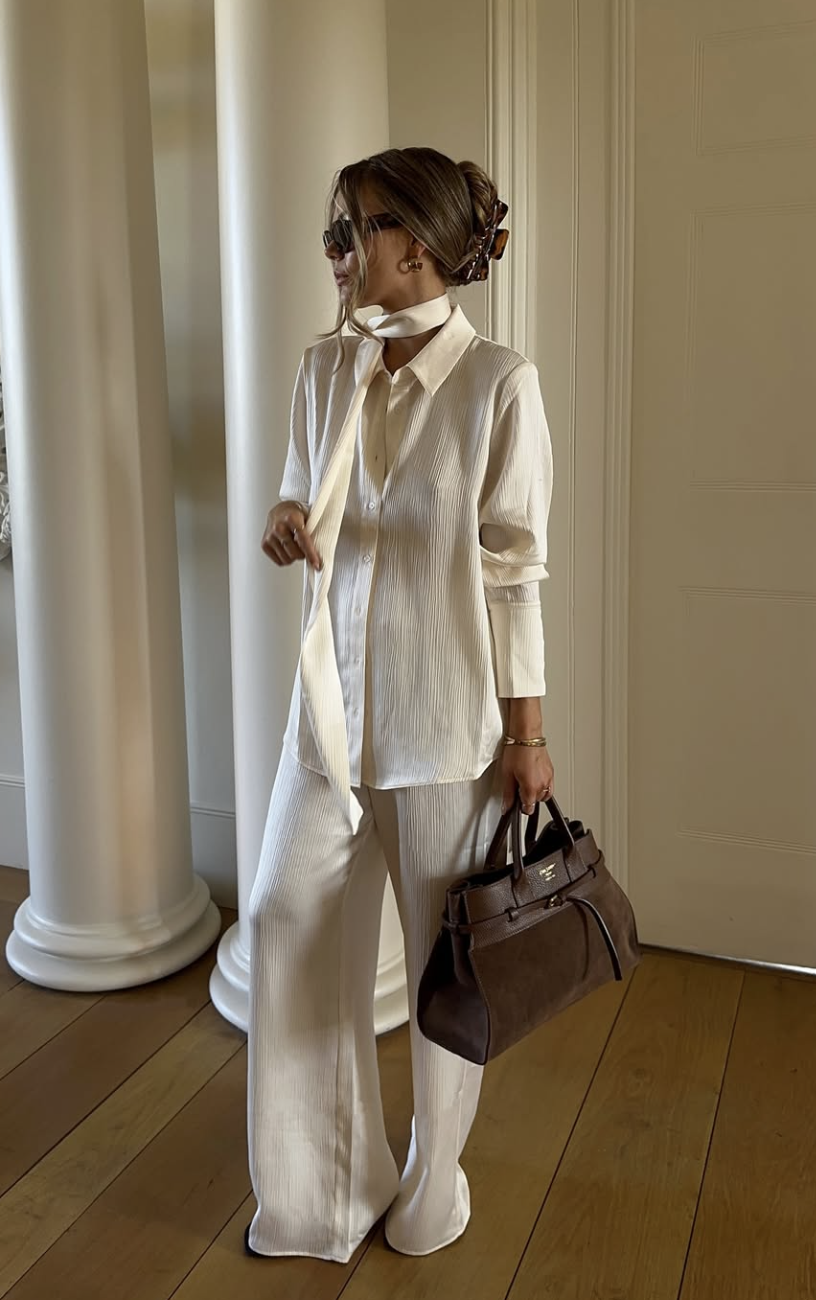 A woman wearing an oversized camel coat over a navy blue sweater and dark-wash wide-leg jeans, styled with a brown leather belt and matching handbag.