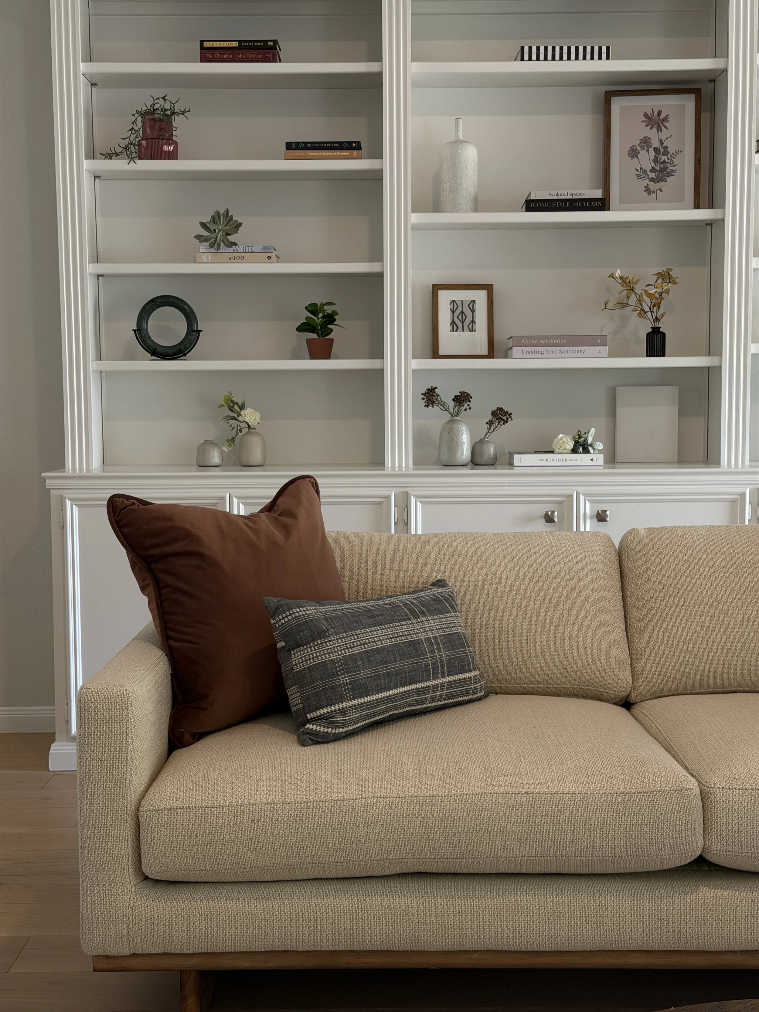 Beige sofa with rust and striped throw pillows in front of white built-in shelves styled with books, plants and framed art
