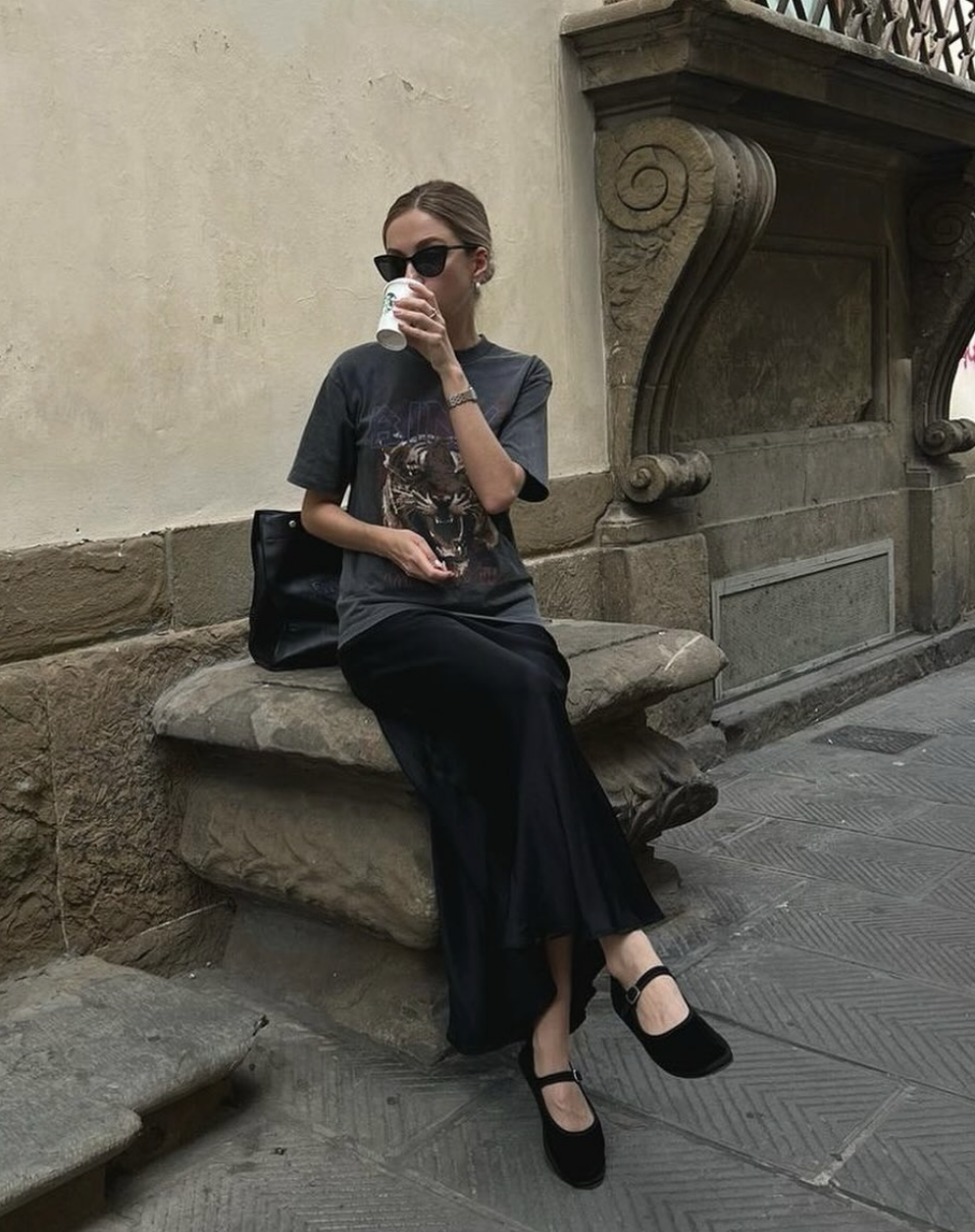 A woman sitting on a stone ledge wearing a charcoal grey graphic t-shirt and a black maxi skirt with black Mary Jane flats for women, holding a coffee cup.