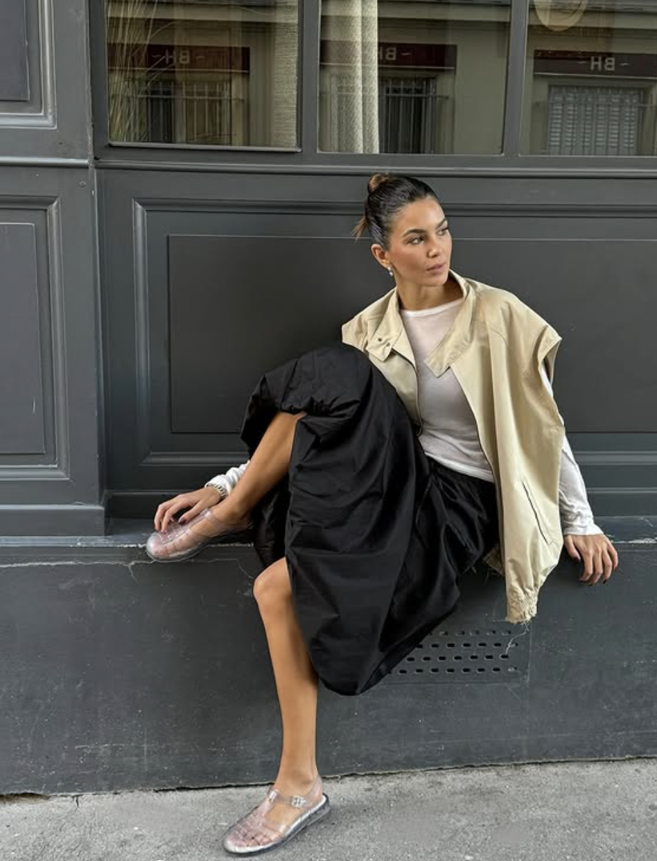 A woman sitting on a stone ledge wearing a voluminous black maxi skirt, a fitted white top, a light cream cropped jacket, and clear translucent jelly Mary Jane flats.