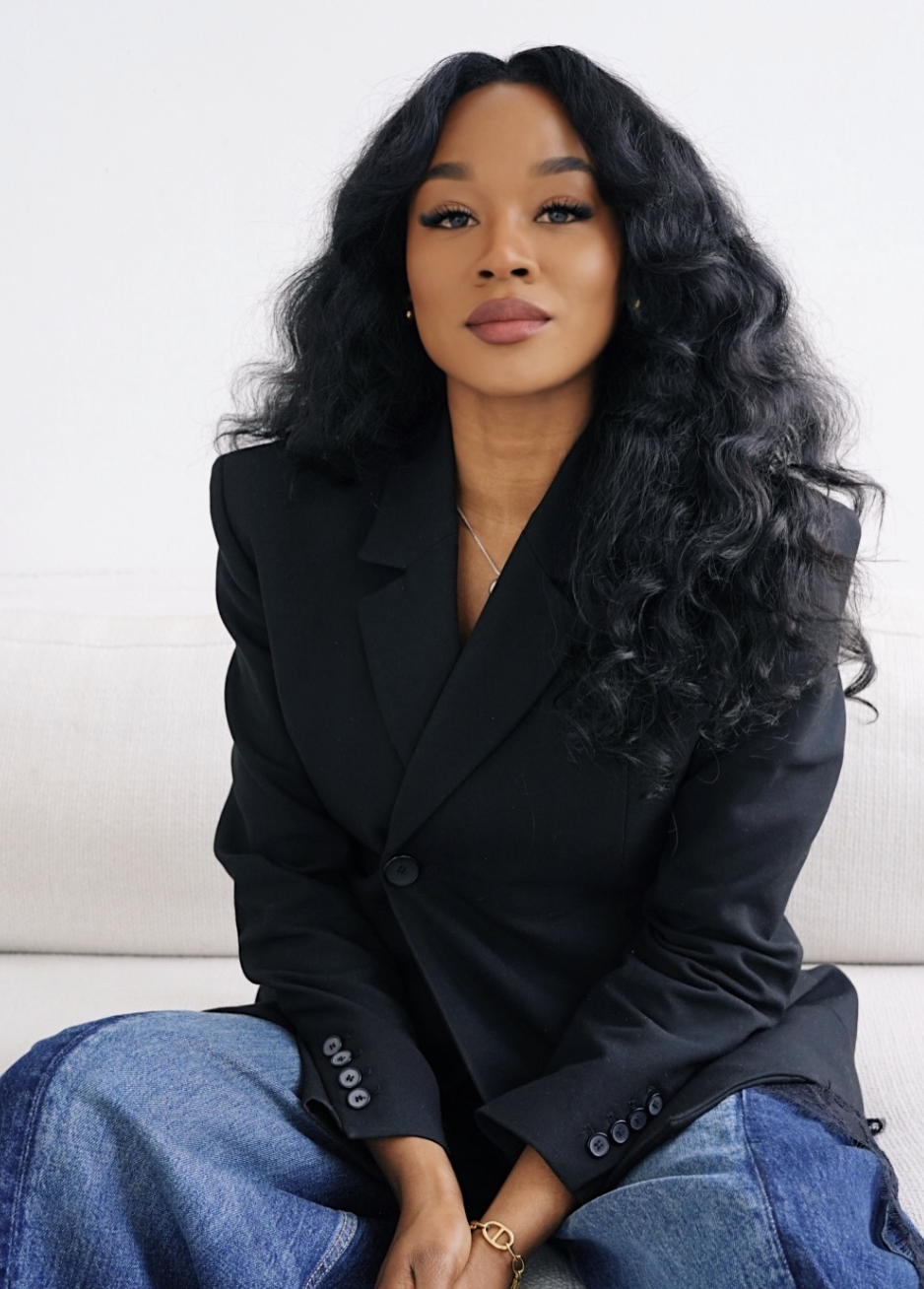A close-up portrait of a woman showcasing voluminous, glossy chunky wand curls in her long black hair, wearing a structured black blazer and a delicate necklace.