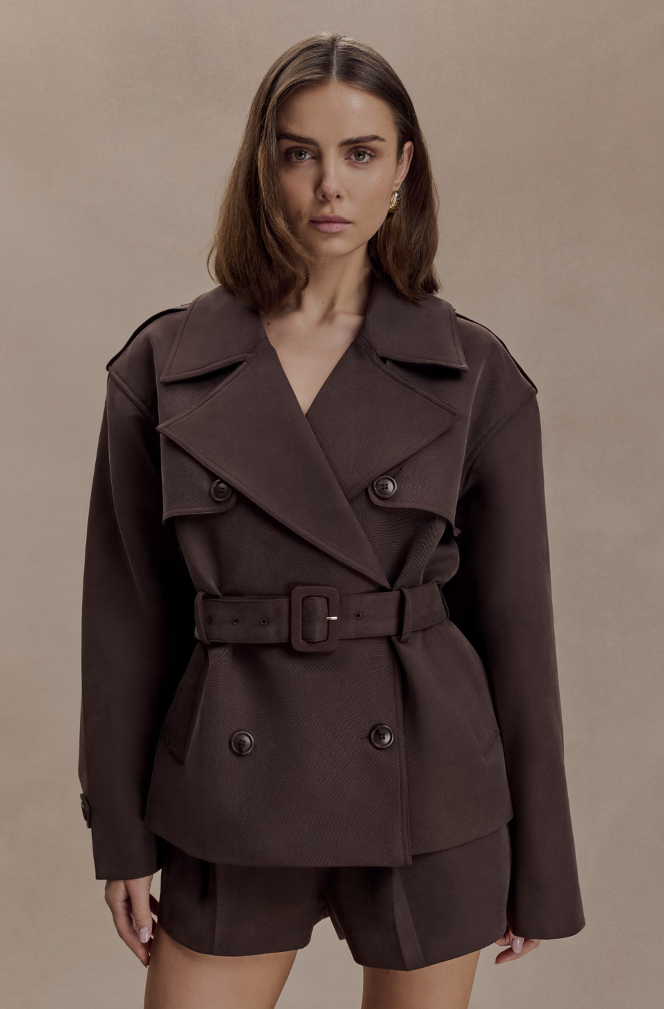 Model wearing the Meshki Alden short trench jacket in chocolate brown, featuring a double-breasted design and a belted waist.