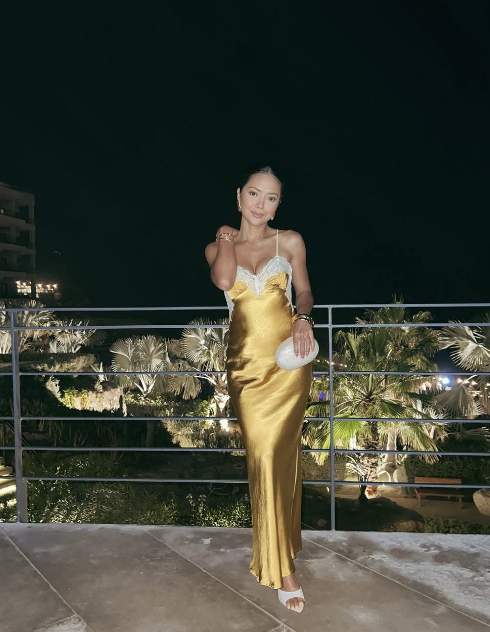A woman styling a glamorous gold satin slip dress with a white lace-trimmed neckline, carrying a white clutch for a rooftop evening look.