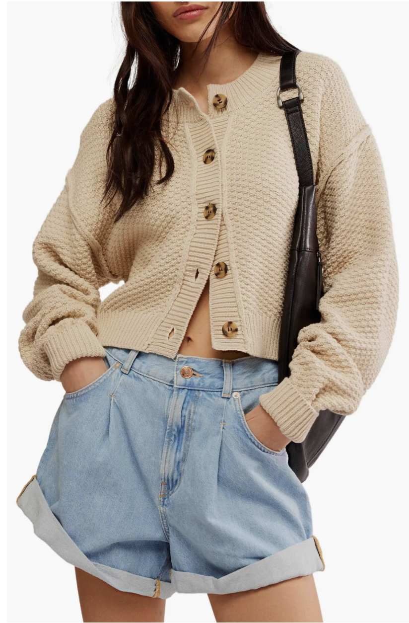 Close-up of a woman wearing a beige Free People Lila crop cardigan with brown buttons and light wash denim shorts.