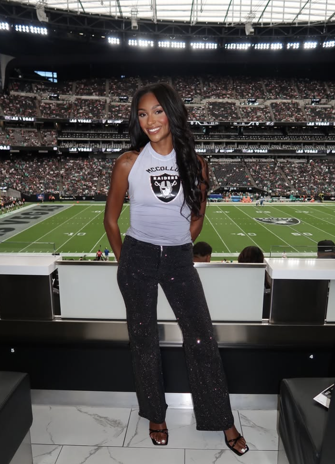 Woman in a football stadium suite wearing a fitted gray Raiders tank, black sparkly wide-leg pants and strappy heels.