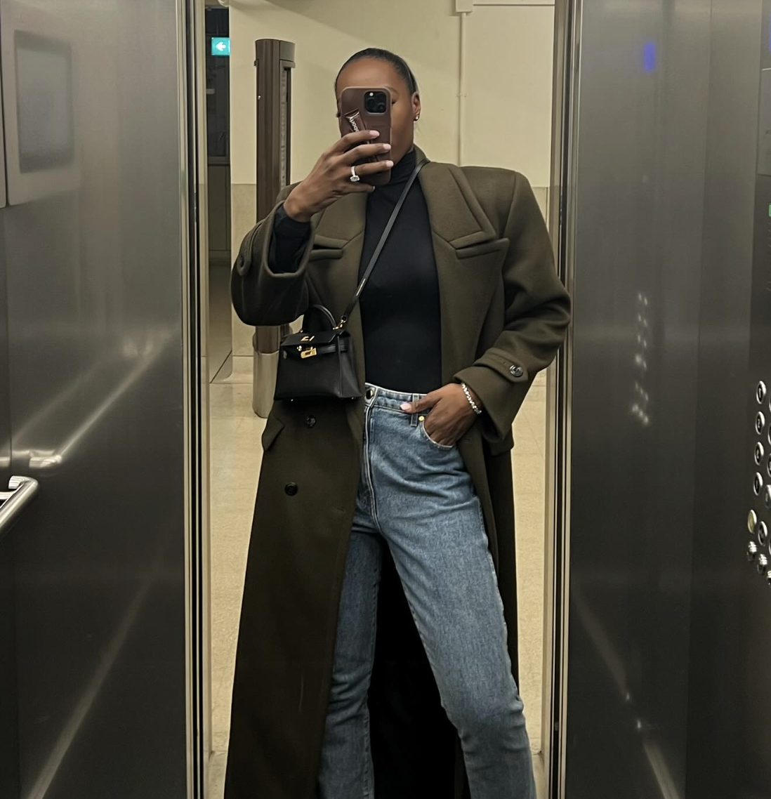 A woman wearing a black fitted turtleneck tucked into blue jeans, layered under a long brown wool coat, styled as a chic everyday look for cooler weather.