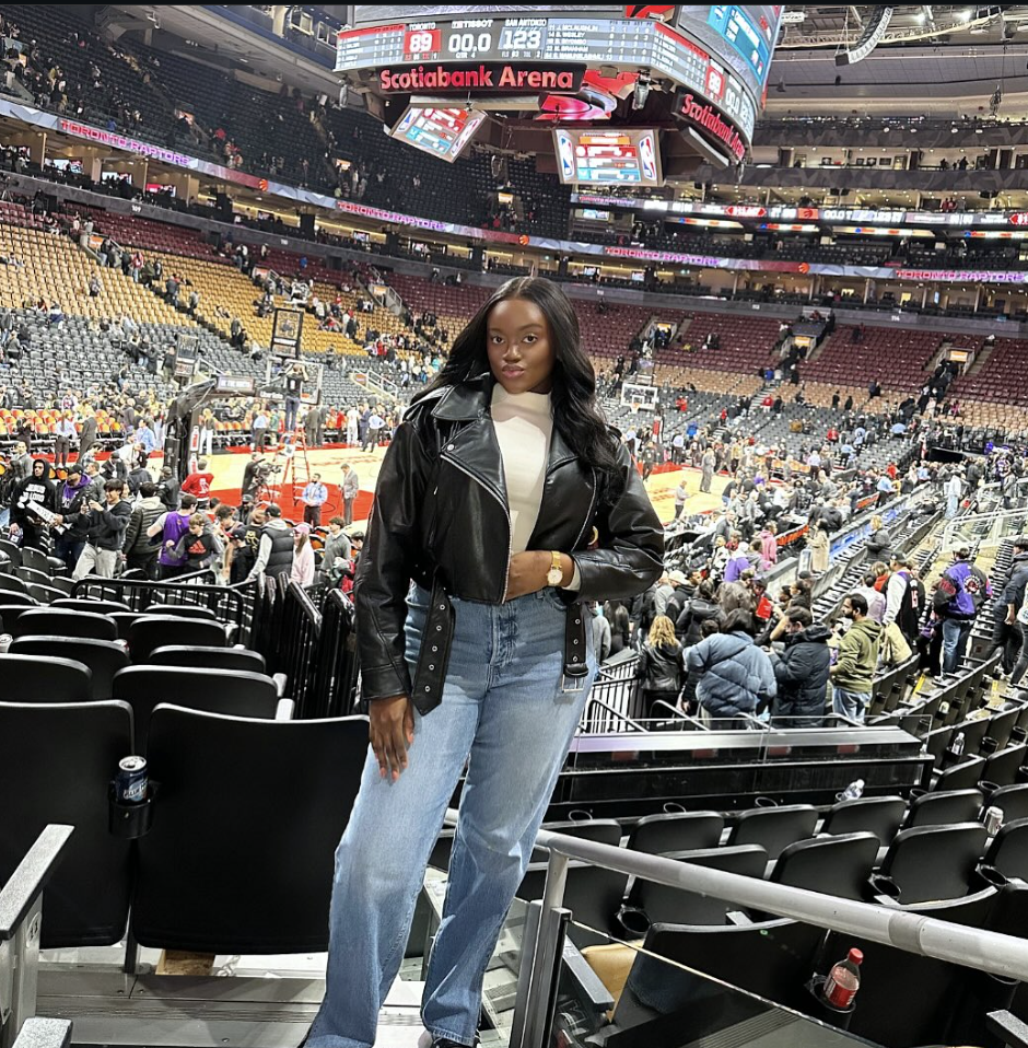 Woman at a basketball arena wearing a black leather bomber, white T-shirt, straight jeans and sneakers.