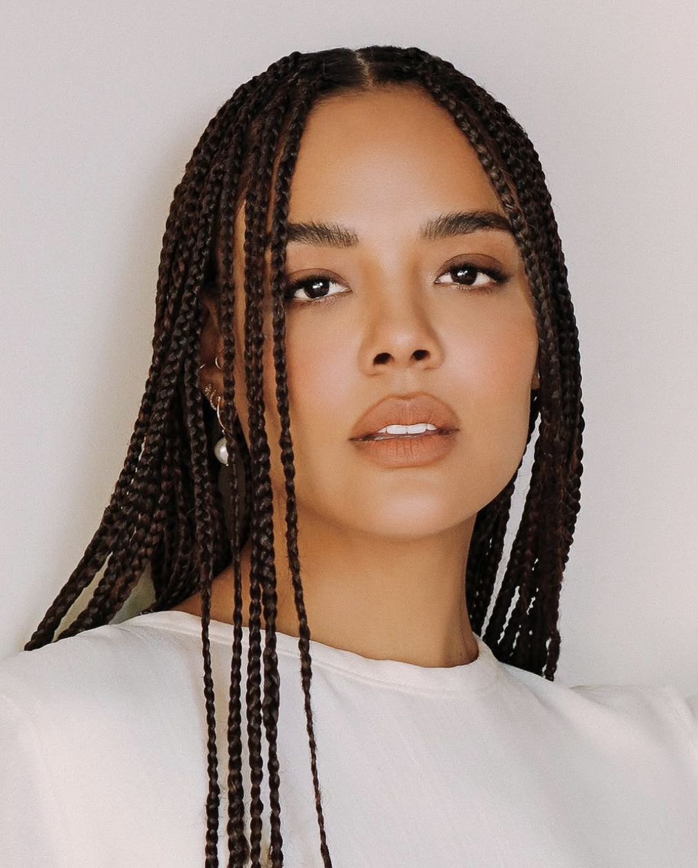 Tessa Thompson with long, dark box braids and a slight side part, wearing a white top, showing how length and vertical lines from braids elongate a round face shape.