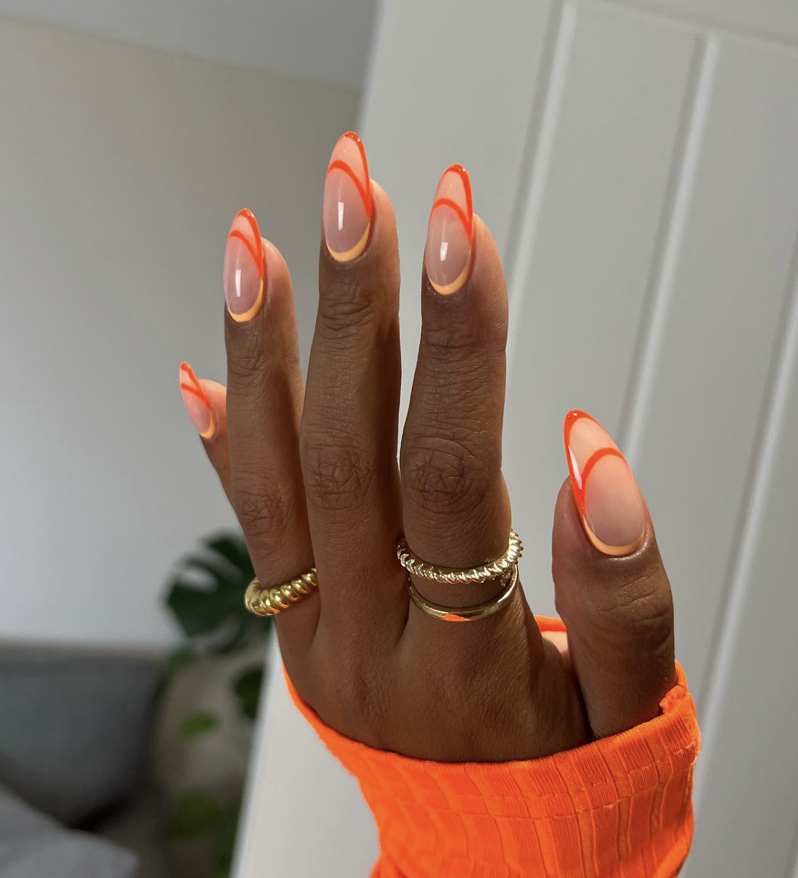 Long, almond-shaped nails with a glossy, semi-sheer base coat featuring a modern, double-layered abstract French tip design in vibrant neon orange and soft pastel yellow waves.