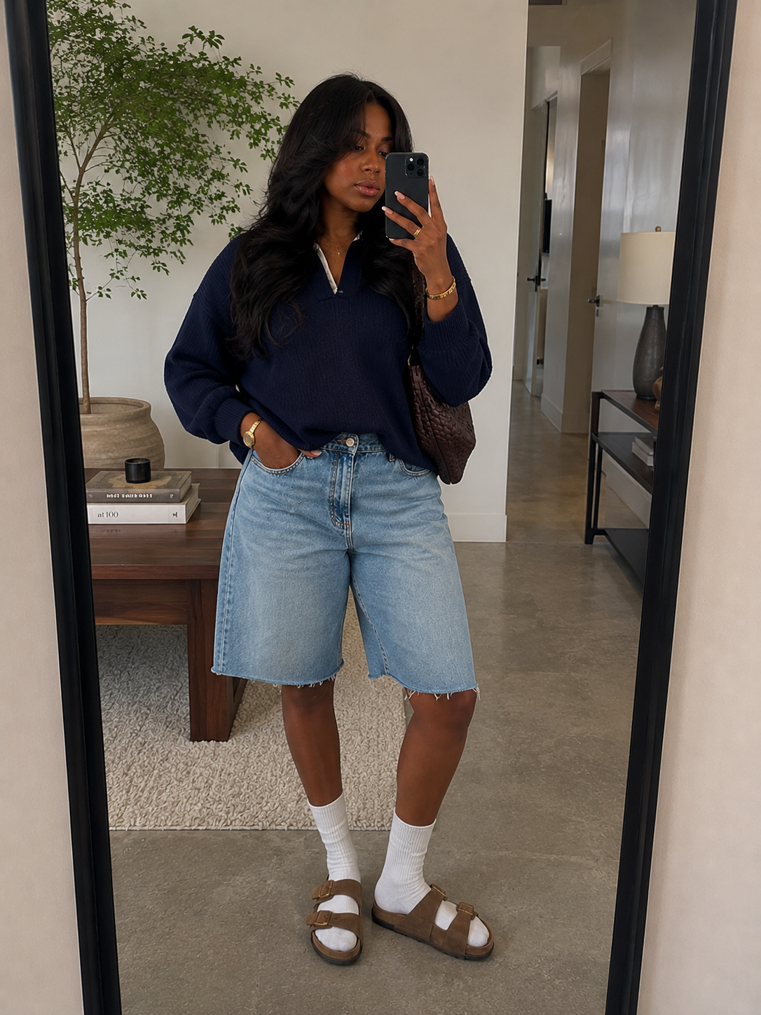 A casual style breakdown featuring a woman from the waist down wearing long, light-wash denim shorts with a raw hem, styled with white crew socks and brown suede Birkenstock Boston clogs.