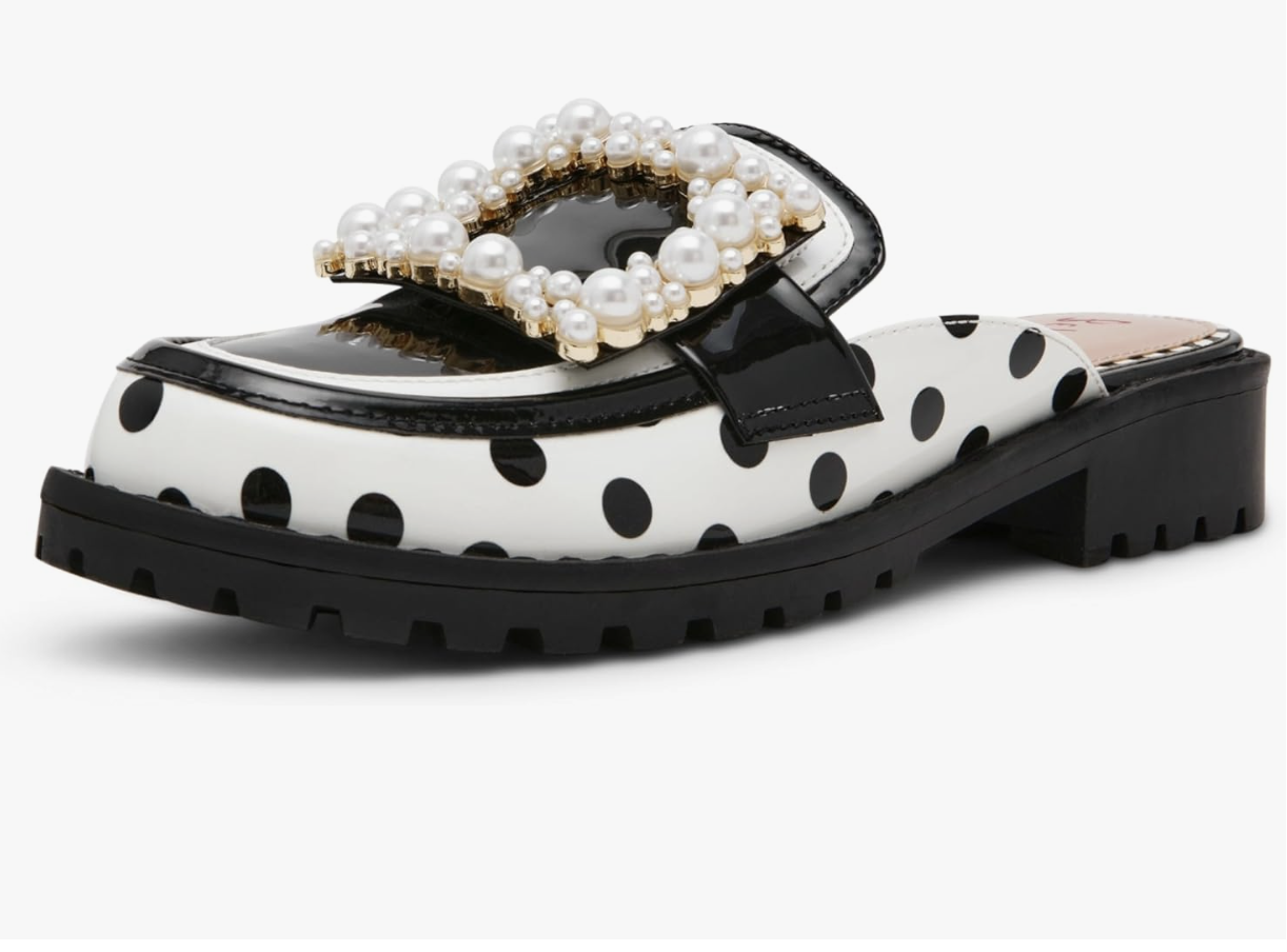 A close-up of the Betsey Johnson Grayce loafer featuring a white leather upper with black polka dots, a chunky black lug sole, and a decorative strap across the top embellished with oversized white pearls.