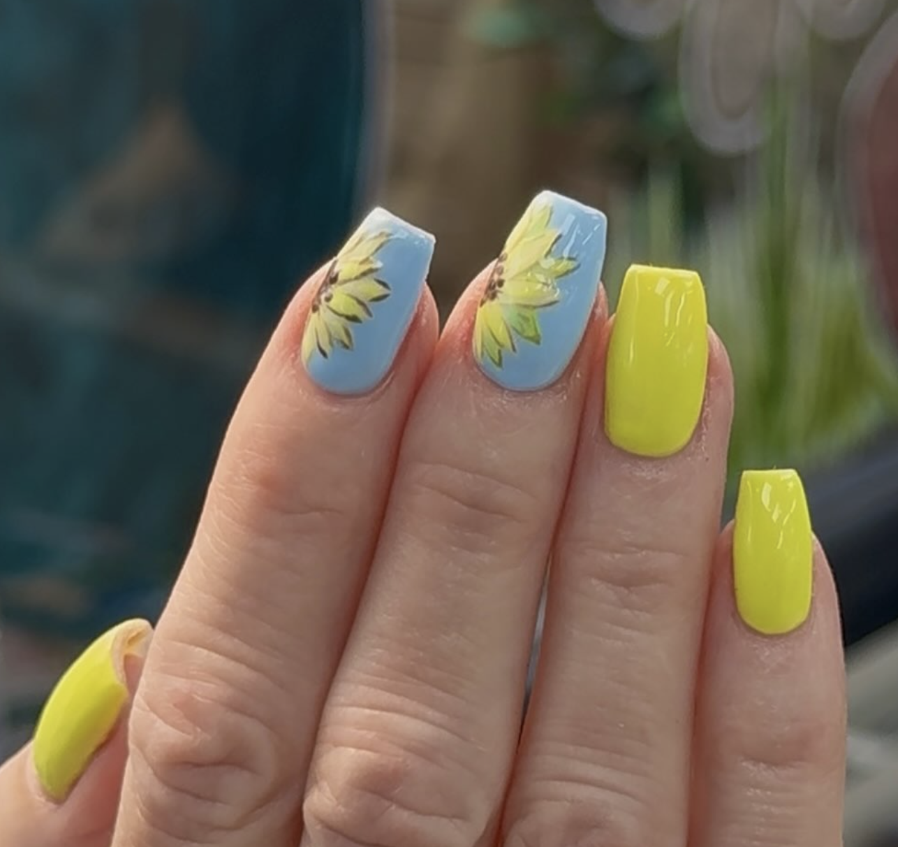 Close-up of almond-shaped nails with a light blue polish, featuring hand-painted sunflowers and a sparkling yellow glitter accent nail.