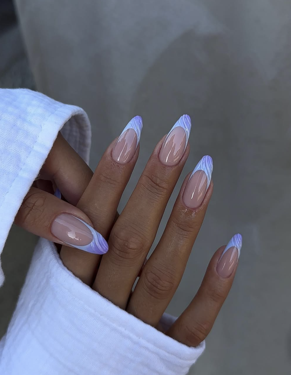 A close-up of long, almond-shaped nails featuring a sheer nude base with a dual-toned lavender and periwinkle blue abstract wave French tip design, styled with a white textured sleeve.