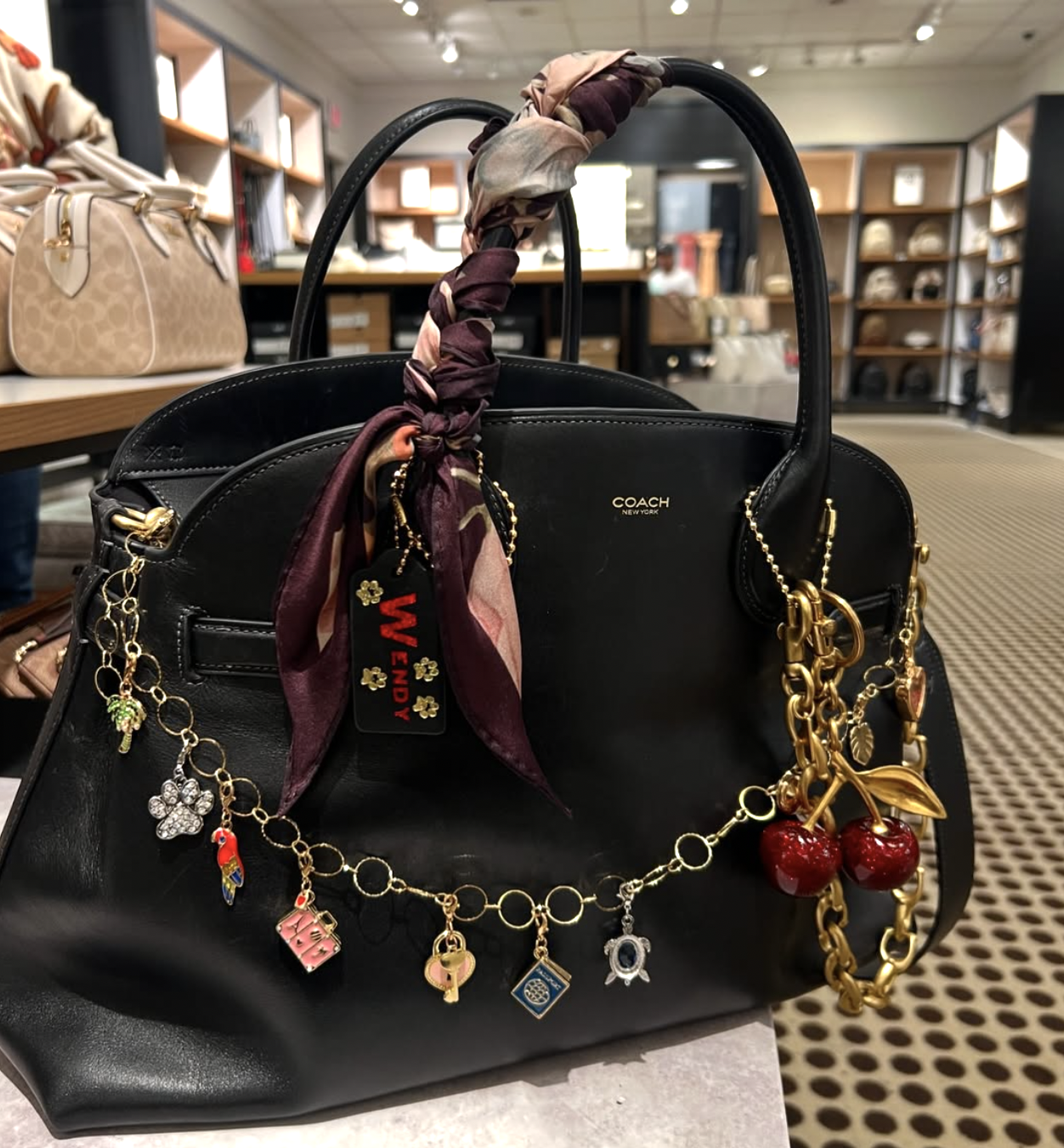 A close-up shot of a black leather Coach handbag decorated with a burgundy and pink patterned silk scarf knotted around the handle, accented by a gold charm chain with travel and nature charms.