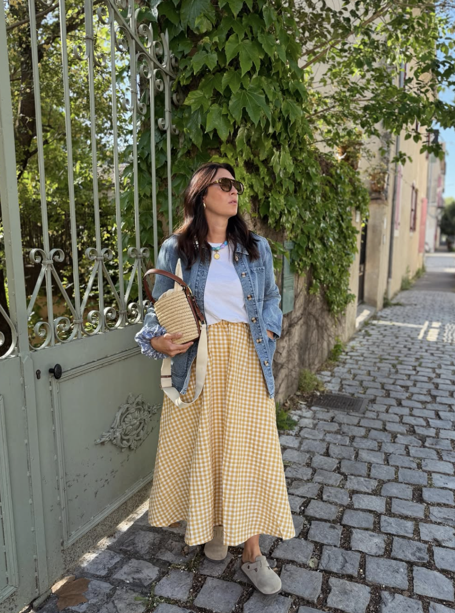 A woman wearing a yellow gingham maxi skirt, a white t-shirt, and a denim jacket, carrying a straw bucket bag.