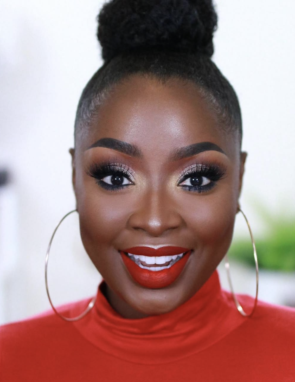 A close-up shot of a woman with a deep skin tone and a high bun, showcasing a Winter color season makeup look with a crisp, saturated true red lipstick and defined eyeliner.