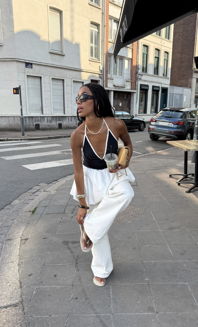 A woman walking on a city street wearing voluminous white wide-leg jeans, a black and white trim halter top, and white thong sandals.