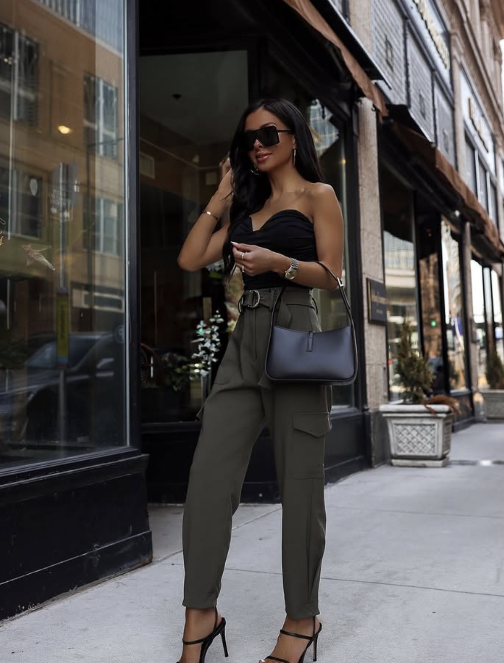 A close-up of a person wearing tapered olive green cargo pants with side pockets, paired with black strappy heeled sandals and carrying a black leather shoulder bag.
