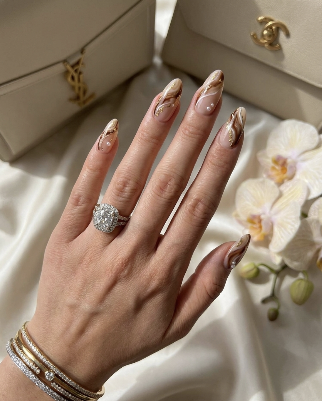 Long, oval-shaped nails with a glossy caramel nude base coat, featuring fine white abstract swirls and delicate gold foil and pearl accents, finished with a soft pearl glaze.