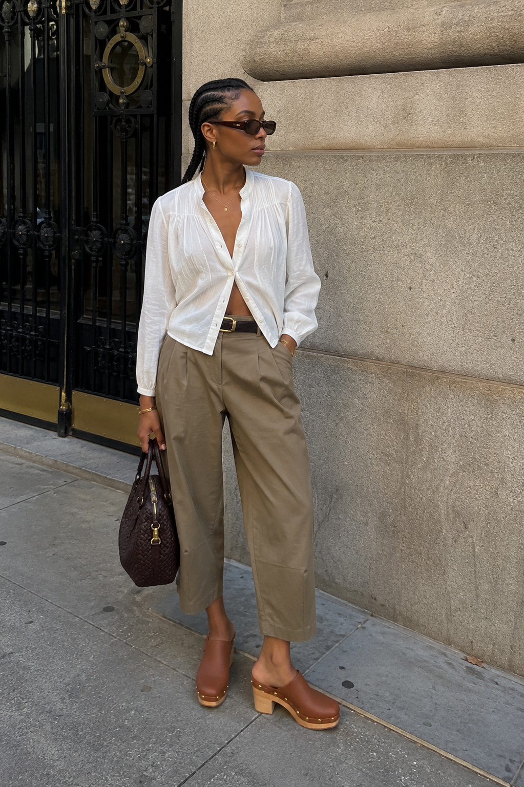 A chic street style outfit featuring a woman wearing high-waisted, pleated cropped trousers in tan, a light white button-down shirt, a dark brown leather belt, and tan platform leather clogs.