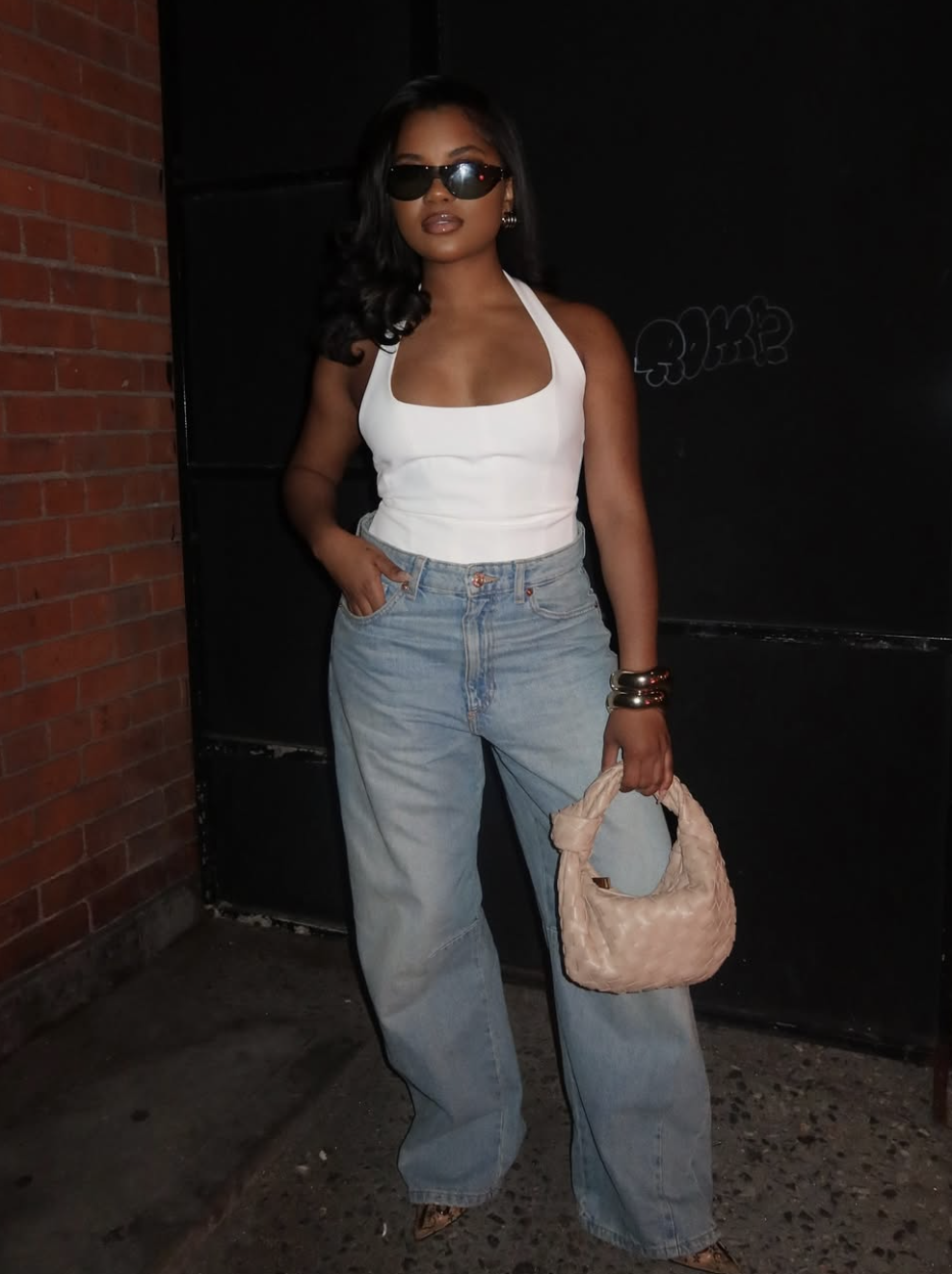 A woman wearing light-wash baggy jeans with a white fitted halter-style ribbed tank top and a woven beige handbag to demonstrate the contrast between a slim top and loose bottom.