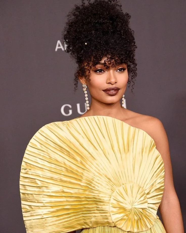 Yara Shahidi wearing a yellow pleated gown and long drop earrings, showcasing a voluminous natural hair high puff that adds vertical height to elongate a round face shape.