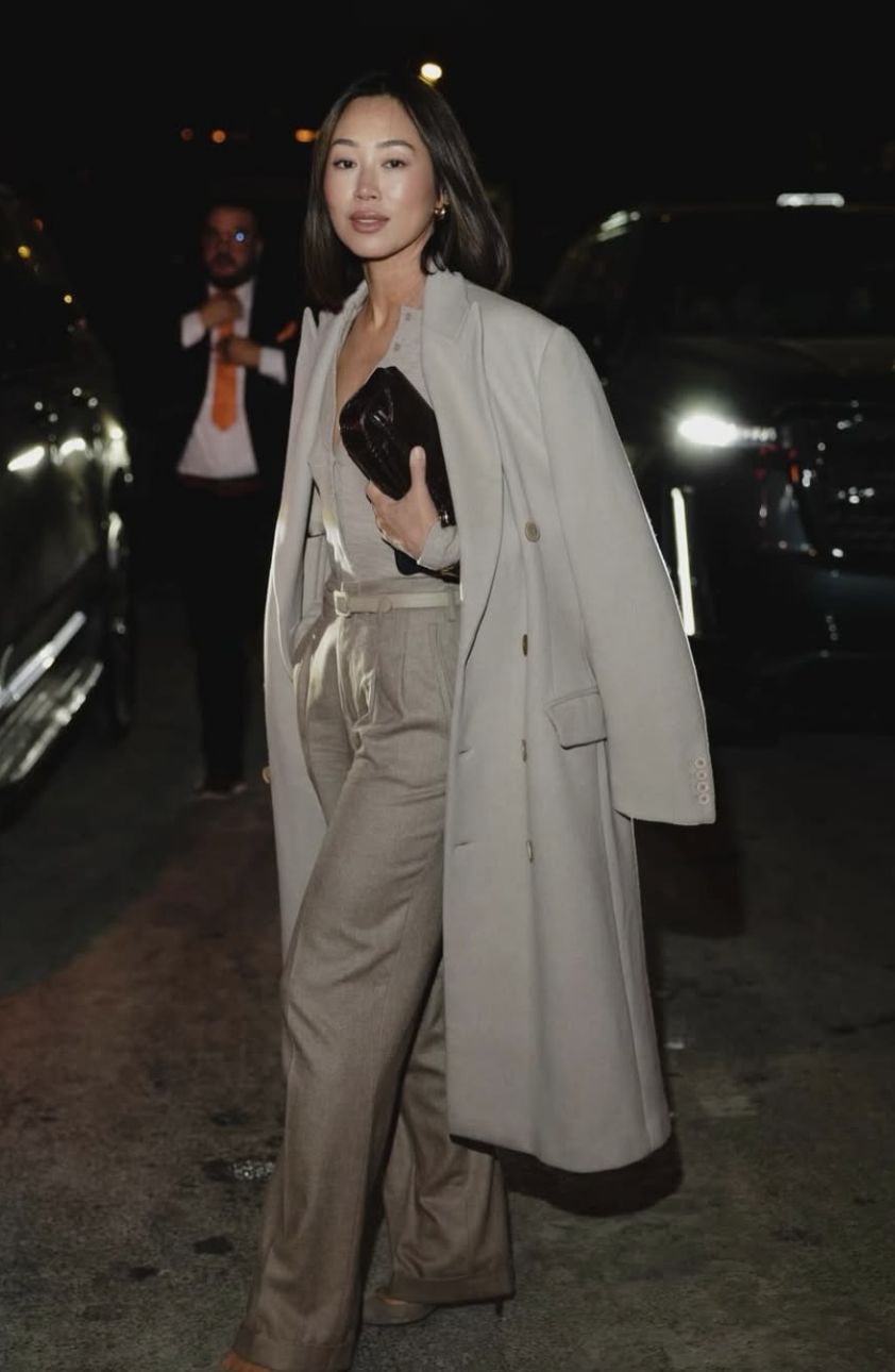 A woman wearing a tan maxi trench coat over a white bodysuit and beige wide-leg trousers, accessorized with a brown woven clutch and animal print heels.