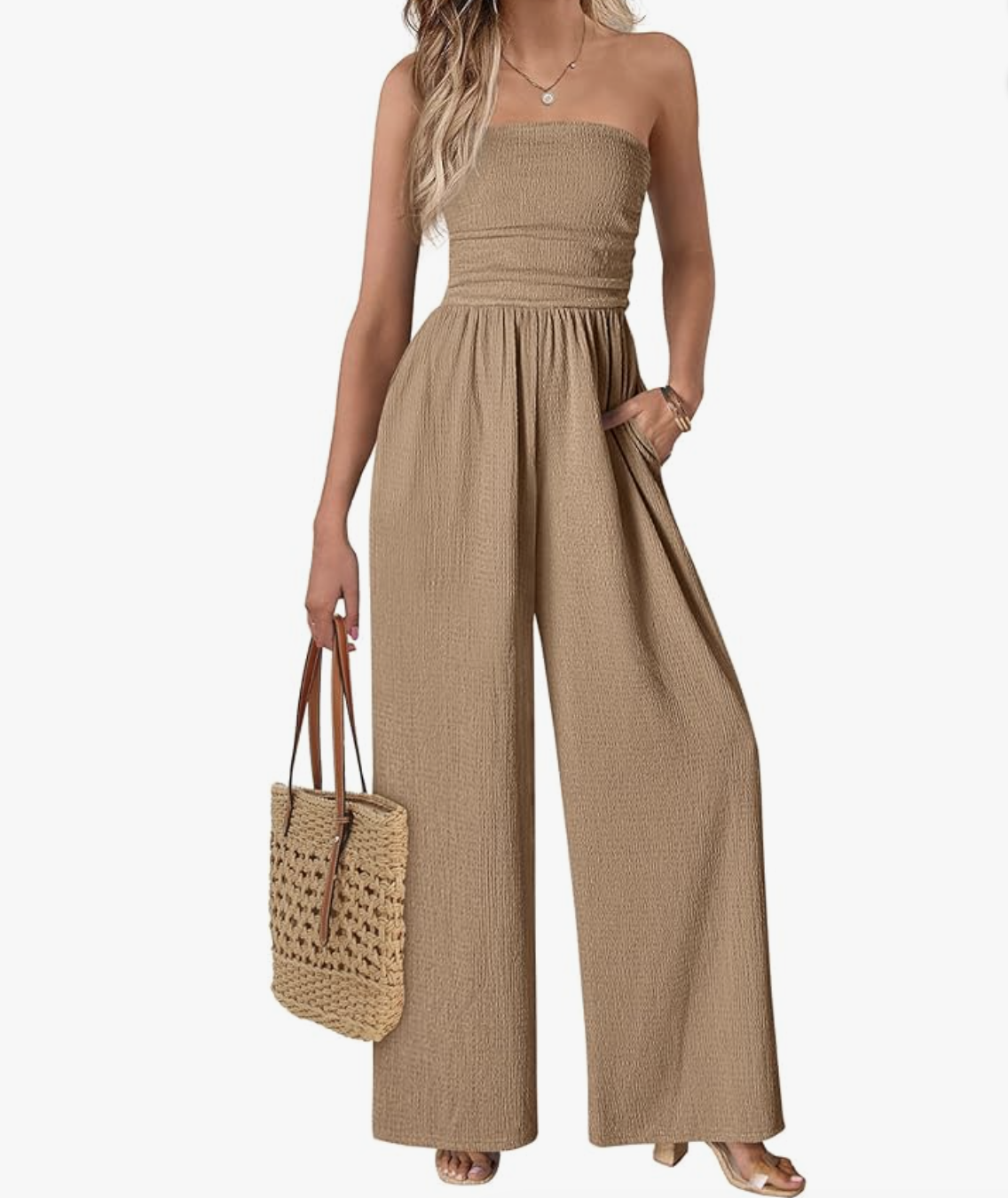 A woman wearing a lightweight, strapless tan jumpsuit featuring a smocked bodice, relaxed wide-leg pants, and side pockets, styled with a woven straw tote bag.