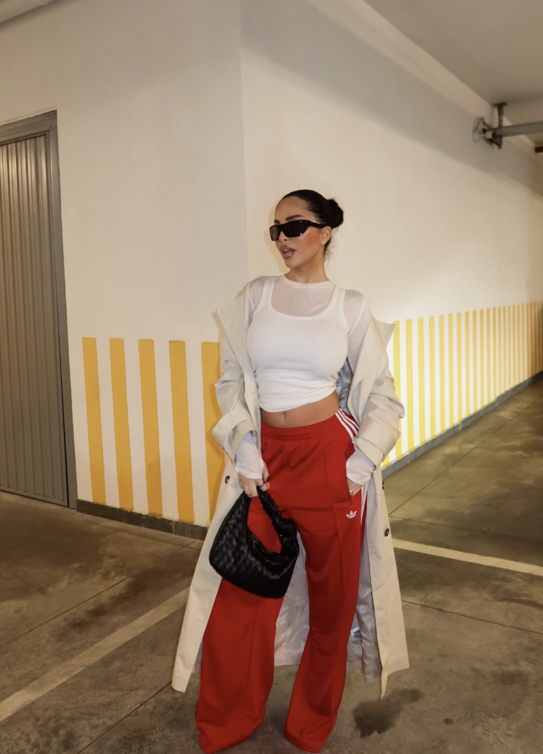 Professional style hack: Red Adidas Firebird track pants paired with a dramatic floor-length black overcoat, a white crop top, and a black woven bag for a high-fashion Downtown Edge aesthetic.
