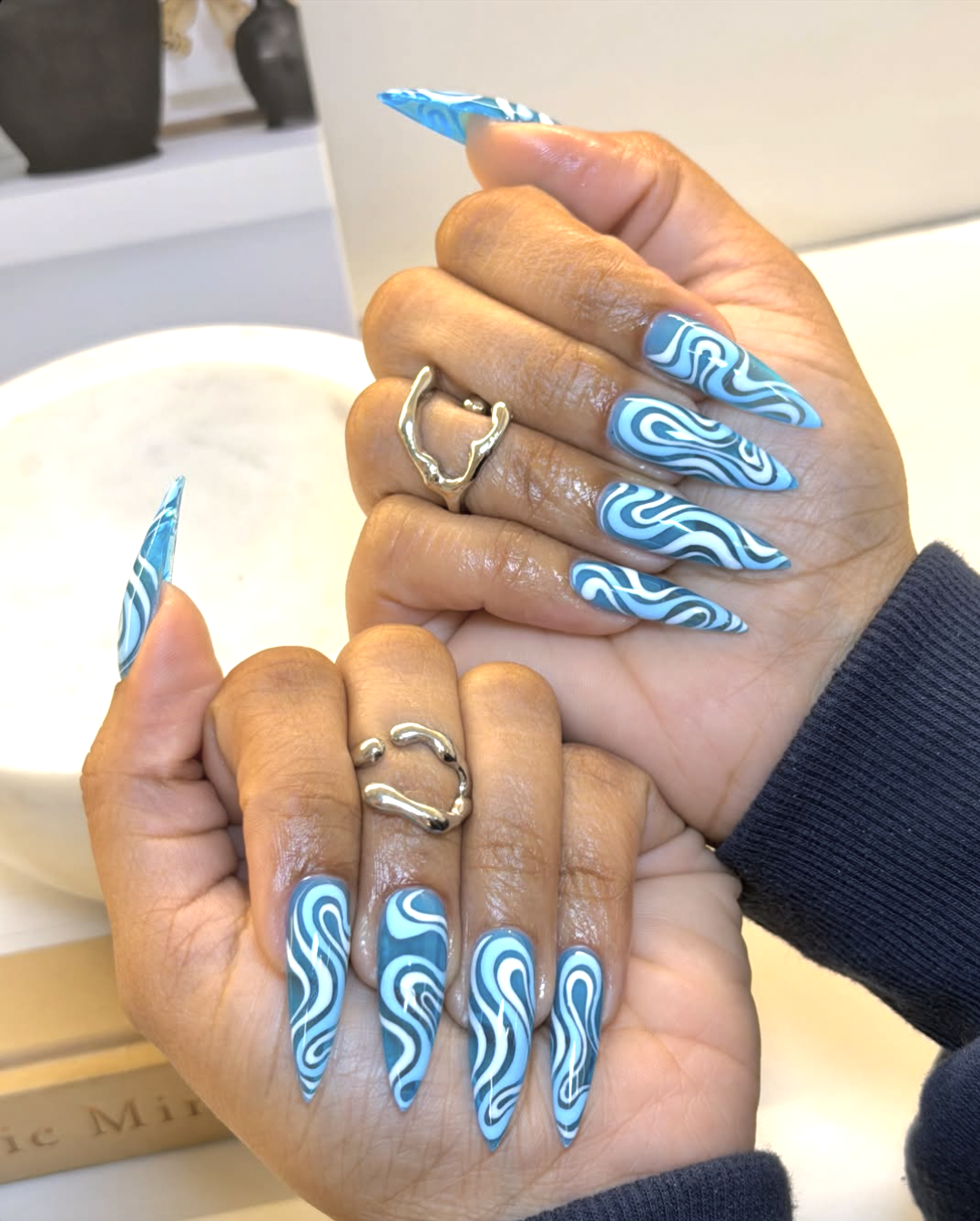 A close-up of long, stiletto-shaped nails featuring an intricate, multi-toned blue and white abstract wave swirl design, heavily accessorized with chunky silver statement rings.