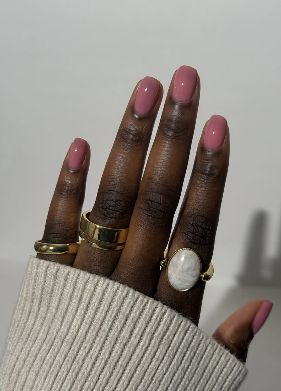 A sheer, milky pink manicure on deep skin tone with gold rings, one of the most versatile summer fingernail colors.