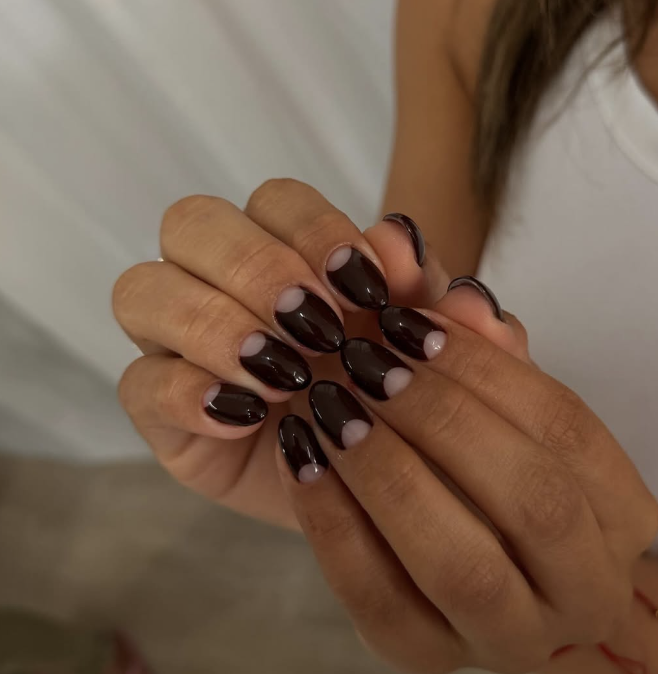 A close-up of glossy deep chocolate brown nails featuring clear half-moon negative space cutouts at the base.