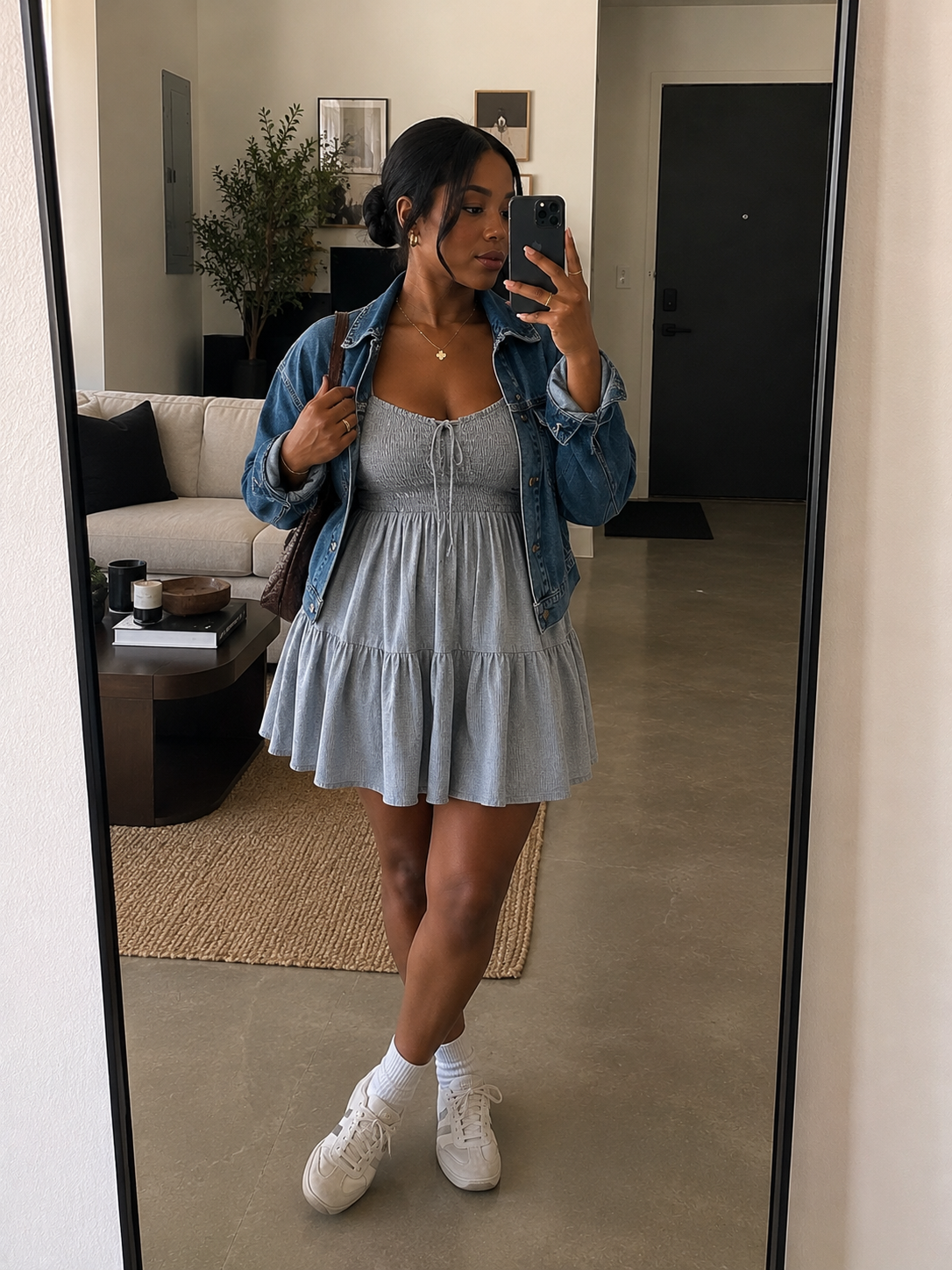 A mirror selfie of a woman styling a casual summer outfit featuring a light blue tiered mini sundress with a smocked bodice, paired with an oversized denim jacket, white crew socks, and white sneakers.