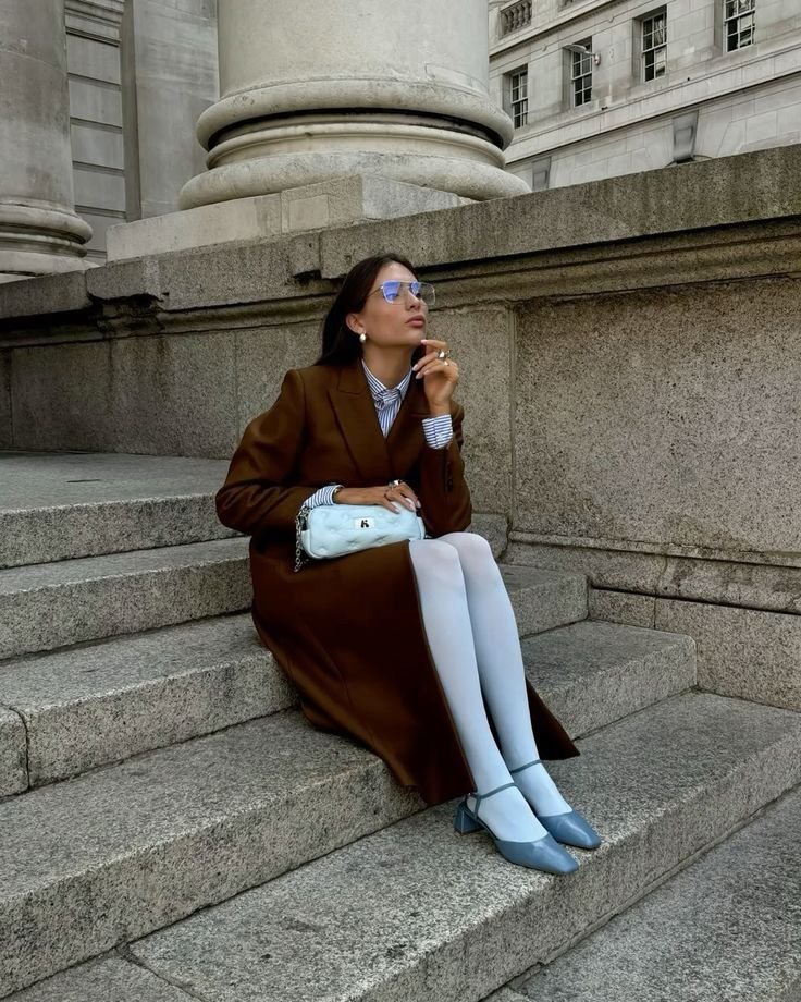 A fashion editor-approved styling for Spring 2026, featuring icy Cool Blue pointed-toe Mary Jane flats paired with opaque white tights and a long chocolate brown coat.
