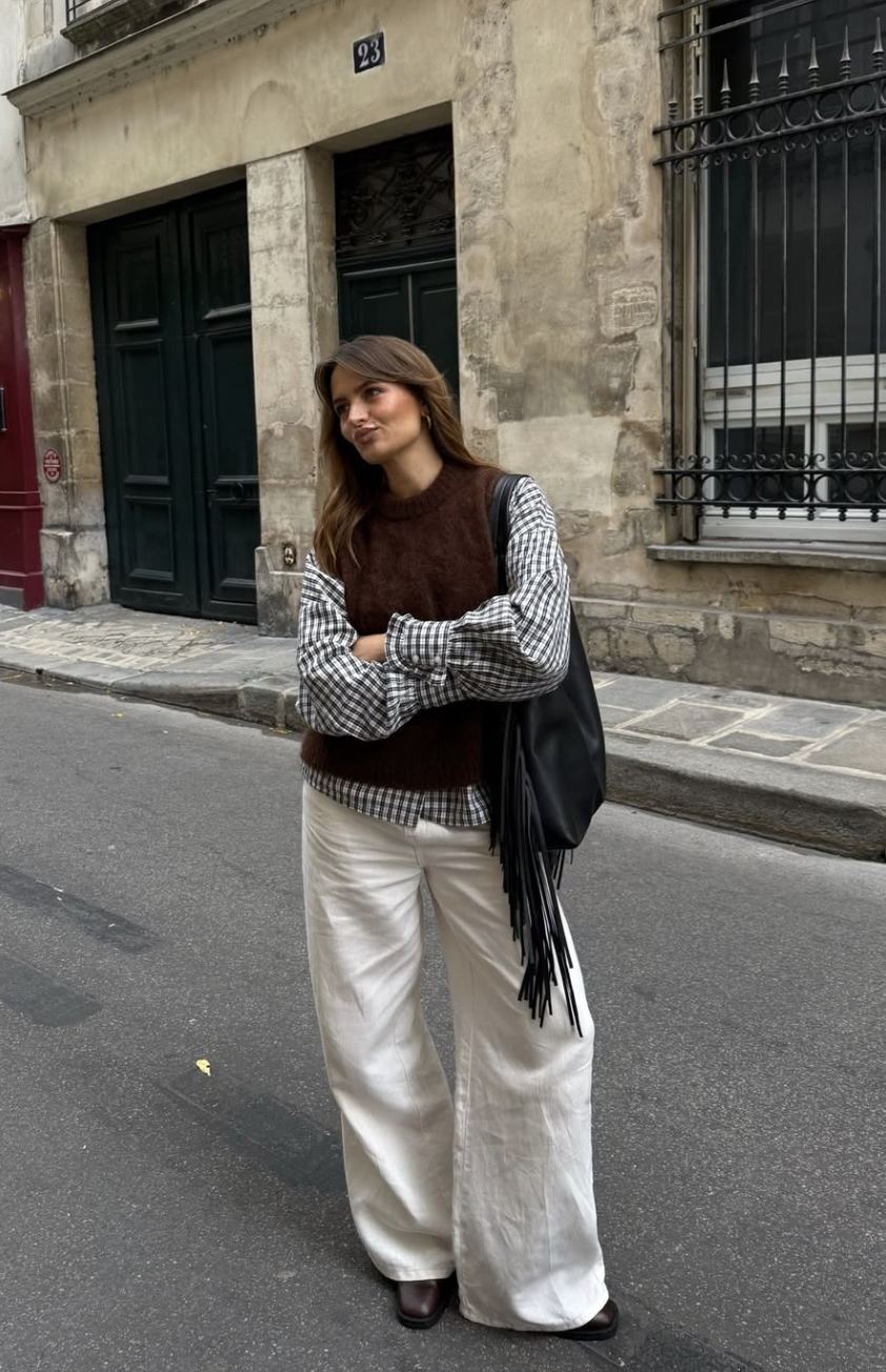 A woman standing on a city street wearing a brown knit vest over a plaid shirt and white trousers, carrying a large black leather tote bag with long fringe tassels.