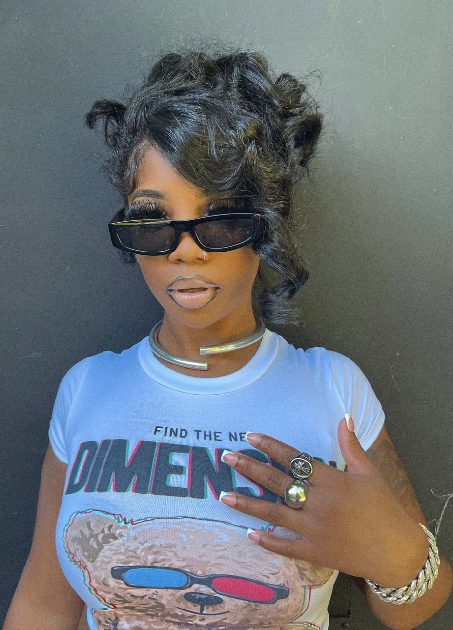 A woman wearing rectangular sunglasses and a blue graphic tee, featuring a high messy bun with side-swept curtain bangs that frame the temples and balance a heart face shape.