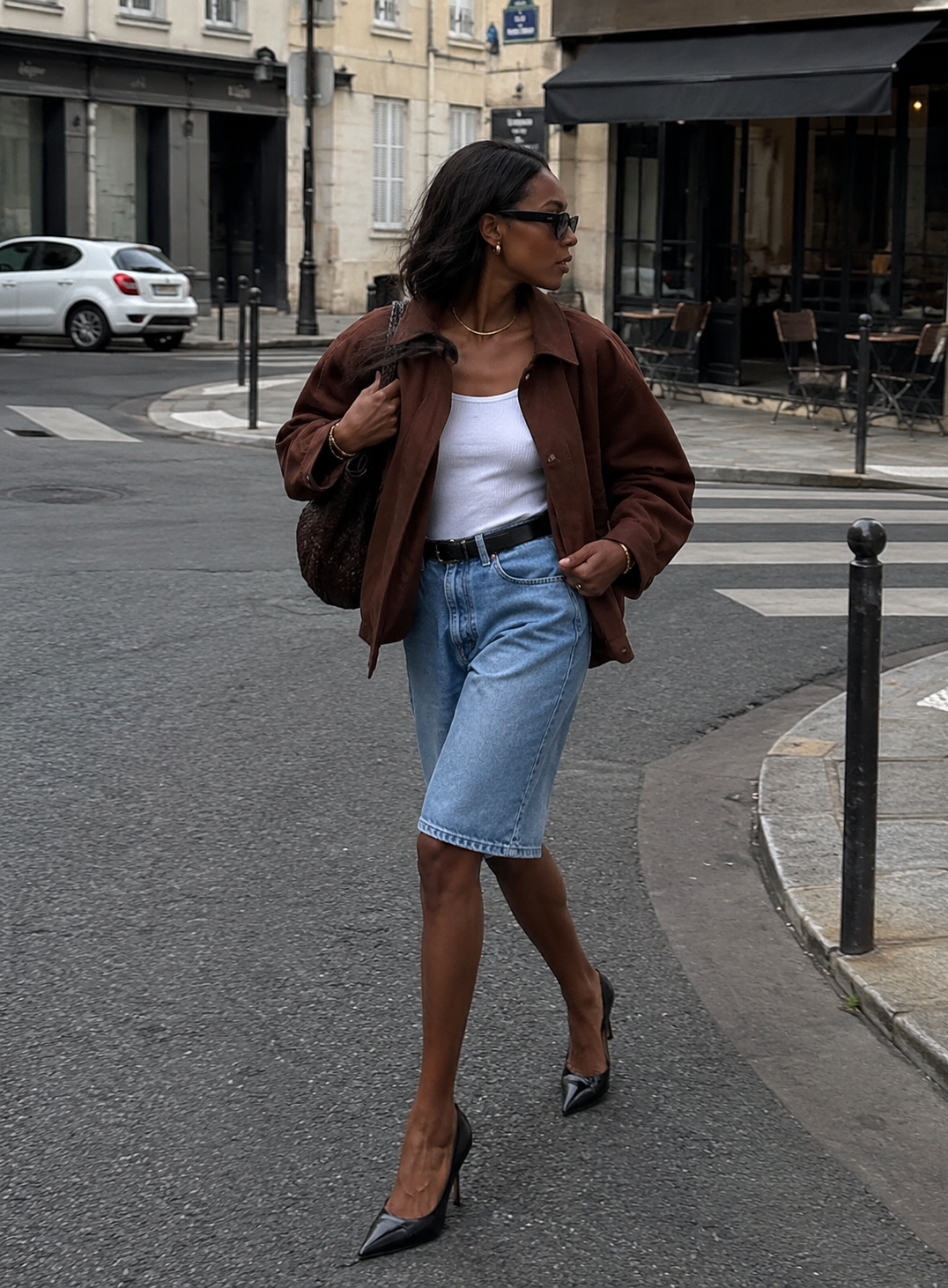A woman walking on a city street wearing long denim Bermuda shorts, a white top, a brown jacket, and black pointed-toe stiletto pumps.